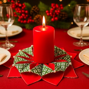 May include: A lit red pillar candle on a decorative red and green fabric centerpiece. The centerpiece has a starburst design with green patterned fabric and white pearl accents. The table setting includes wine glasses and plates on a red tablecloth.