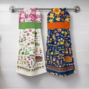 以下が含まれることがあります： 2つの吊り下げ式キッチンタオル。1つはピンクの花柄と緑色のバンド、もう1つは青色の花柄とオレンジ色のバンドです。どちらも庭をテーマにしたイラストと「herbs」と「my garden」の文字が特徴です。