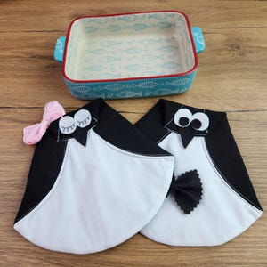 Puede incluir: Una fuente rectangular de cerámica para hornear con borde azul y estampado de peces, junto a dos agarraderas con forma de pingüino. Las agarraderas son negras y blancas con lazos, uno rosa y otro negro.