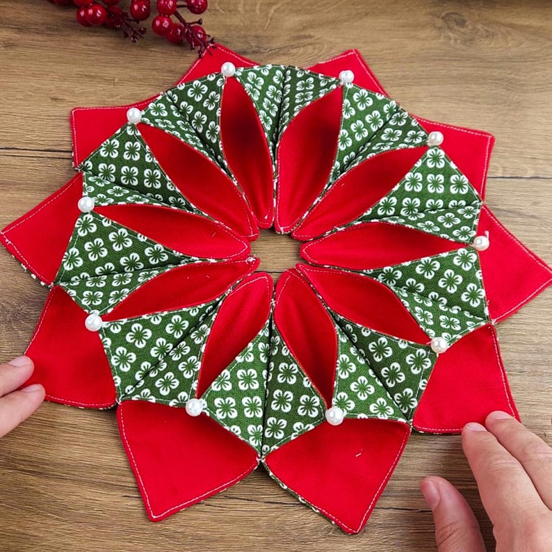 May include: A handmade, star-shaped fabric item with a red and green floral pattern. The item is made of quilted fabric and has white pearl accents. The design is symmetrical and appears to be a decorative item.