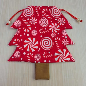 May include: A red Christmas tree-shaped drawstring bag with a white candy cane and swirl pattern. The bag has a brown rectangular base and red drawstrings with white pearl-like beads.