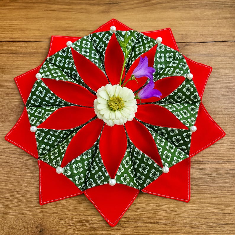 May include: A decorative, star-shaped placemat with a red base and a floral design. The center features red petals alternating with green fabric patterned with white flowers. White pearls and fresh flowers add detail.