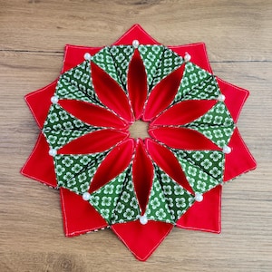 May include: A handmade, star-shaped decorative item with a red base and a central floral design. The floral design features green fabric with white flower patterns and red inner petals. White pearl accents are placed around the edges.