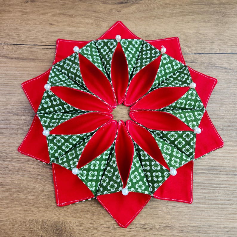 May include: A handmade, star-shaped decorative item with a red base and a central floral design. The floral design features green fabric with white flower patterns and red inner petals. White pearl accents are placed around the edges.