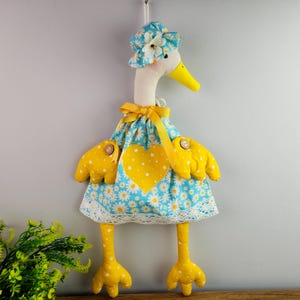 May include: A handmade goose decoration, featuring a blue dress with a daisy pattern and a yellow heart. The goose has yellow wings, legs, and a matching hat. It is designed to be hung on a wall.