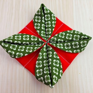 May include: A handmade fabric item with a red square base and four green, leaf-shaped accents. The green fabric features a white floral pattern. The item is likely a decorative element or a component of a larger craft project.