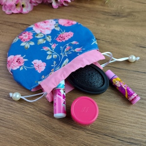 Puede incluir: Una bolsa azul floral con cordón y ribete rosa, que contiene un cepillo para el pelo negro, dos bálsamos labiales y un pequeño recipiente rosa. El estampado floral de la bolsa presenta rosas rosas y hojas verdes. Los artículos están dispuestos sobre una superficie de madera.