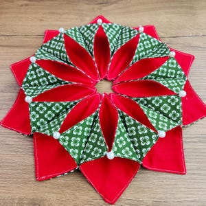 May include: A handmade, star-shaped Christmas ornament. The ornament features a red base with green and white floral patterned fabric accents. Small white beads are placed at the points where the fabrics meet. The ornament is designed for holiday decor.