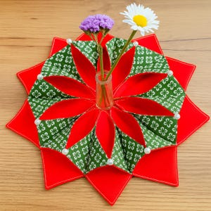 May include: A red, star-shaped table centerpiece with a green and white patterned design. The centerpiece holds a small glass vase with purple and white flowers. White pearl accents are placed around the edges.