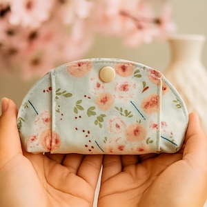 May include: A small, light blue floral print pouch with a rounded top, held in open hands. The pouch features a pattern of pink roses and green leaves, secured with a beige button closure. The background is blurred, with hints of pink flowers and a white vase.