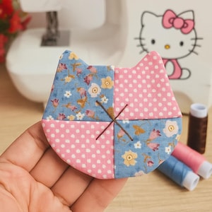 May include: A handmade cat-shaped fabric patch. The patch is divided into four sections: two pink polka-dot and two blue floral print. Brown thread forms an 'X' across the center. Sewing supplies and a Hello Kitty image are in the background.