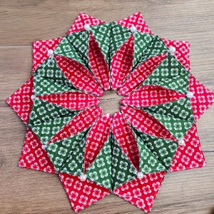 May include: A handmade, star-shaped coaster with red and green floral patterned fabric. The coaster is segmented into triangular sections, with white pearl accents at the points. The design is symmetrical and festive.