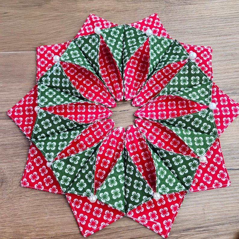 May include: A handmade, star-shaped coaster with red and green floral patterned fabric. The coaster is segmented into triangular sections, with white pearl accents at the points. The design is symmetrical and festive.