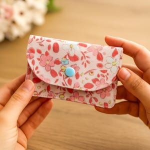 以下が含まれることがあります： ピンクと赤の色合いの小さな花柄の財布で、ボタン留めが付いています。財布は両手で持たれており、そのコンパクトなサイズとデザインを示しています。花柄にはピンクの花と緑の葉が含まれています。