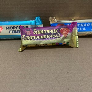 May include: Three wrapped bars, possibly food items, in a cardboard box. The wrappers are in shades of blue, gold, and purple, with text in a non-English script. The bars are arranged side by side, suggesting a product display.