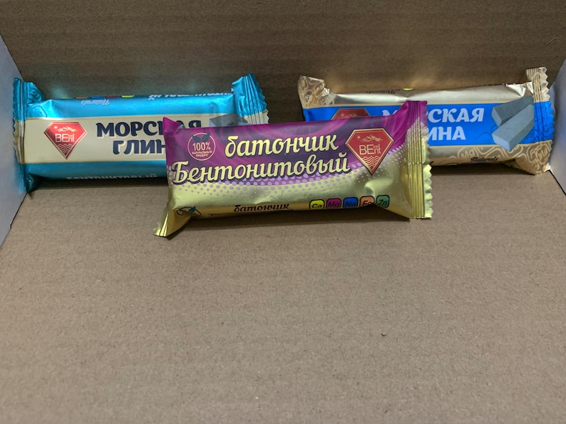 May include: Three wrapped bars, possibly food items, in a cardboard box. The wrappers are in shades of blue, gold, and purple, with text in a non-English script. The bars are arranged side by side, suggesting a product display.