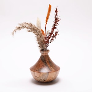 May include: A wooden vase with a unique shape, holding dried flowers in shades of brown, orange, and white. The vase has a wide base and a narrow neck, showcasing the natural wood grain. A decorative piece for home decor.