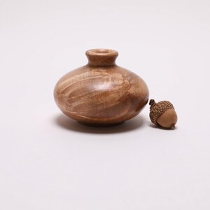 May include: A small, round wooden vase with a narrow neck and a light brown finish. Next to it is a small acorn. The vase and acorn are set against a white background.