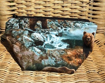 May include: A zippered pouch featuring a detailed landscape with a bear, waterfall, and rocky terrain. The pouch has a black zipper and is set against a woven wicker background.