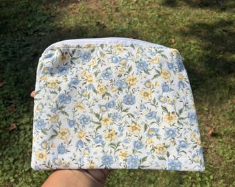 May include: A floral print cosmetic bag with a white background and a pattern of blue and yellow flowers. The bag has a zipper closure and is held against a background of green grass.