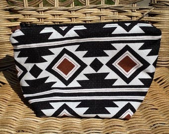 May include: A black, white, and brown patterned cosmetic bag. The bag features a geometric design with diamond and triangle shapes. The bag is sitting on a woven wicker chair.