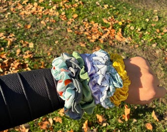 May include: A collection of colorful scrunchies in various patterns and colors, including floral, geometric, and solid yellow. The scrunchies are made of fabric and are worn on the wrist. The background is a grassy area with fallen leaves.
