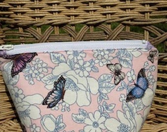 May include: A pink zippered pouch with a floral and butterfly pattern. The design features white flowers and blue and purple butterflies. The pouch has a white zipper and is set against a woven background.
