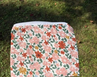 May include: A floral print pouch with a white zipper. The pouch features a pattern of pink, orange, and yellow flowers with green leaves on a white background. The pouch is held against a background of green grass.