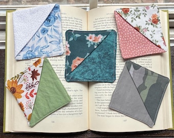 May include: Five triangular fabric corner bookmarks resting on an open book. The bookmarks feature various patterns including blue roses, teal floral, pink floral, autumn leaves with sunflowers, and a grey camouflage print with solid fabric accents.