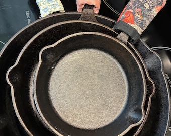 May include: Three black cast iron skillets nested together, each featuring a fabric handle cover. The covers display distinct patterns: one with blue and yellow flowers, one with orange and pink floral motifs, and one with a dark rooster print.