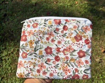 May include: A floral patterned pouch with a white zipper. The pouch features a white background with a repeating pattern of red, orange, and pink flowers with green leaves. The pouch is rectangular and appears to be made of fabric.