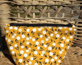 May include: A yellow floral print cosmetic bag with a black zipper. The bag features a pattern of white flowers and black stems on a yellow background. The bag is sitting on a wicker chair.