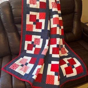 May include: A red, white, and blue patchwork quilt with a navy blue border. The quilt is folded and draped over a brown leather couch.