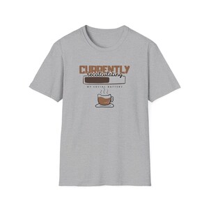 May include: A heather gray short-sleeve t-shirt featuring a graphic with the text "CURRENTLY recalculating" above a loading bar and the text "MY SOCIAL BATTERY" followed by an illustration of a steaming cup of coffee on a saucer.