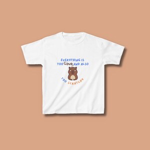 May include: A white short-sleeve t-shirt featuring a brown cartoon capybara graphic in the center. Above the capybara, the text reads "EVERYTHING IS TOO LOUD" in blue, and below it, the text reads "AND ALSO TOO SCRATCHY" in orange letters.