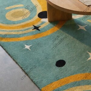 May include: A teal rug with a celestial design featuring yellow and cream-colored concentric circles, black dots, and white stars. The rug is partially visible, with a wooden table leg and tabletop in the background. The text "rugs craftsshop" is printed on the rug.