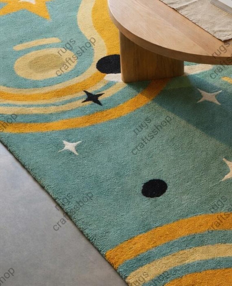 May include: A teal rug with a celestial design featuring yellow and cream-colored concentric circles, black dots, and white stars. The rug is partially visible, with a wooden table leg and tabletop in the background. The text "rugs craftsshop" is printed on the rug.