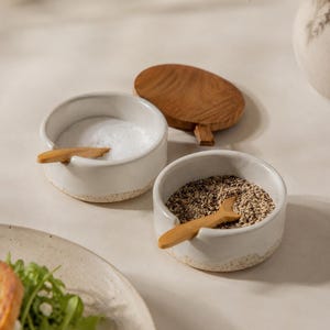 Op de afbeelding: Twee ronde witte keramische kommen met gespikkelde bodems, elk met een kleine houten lepel. De ene kom is gevuld met wit zout, de andere met zwarte peper. Een rond houten deksel met een klein handvat ligt naast de zoutkom.