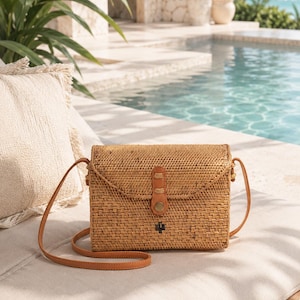 A woven straw handbag with a leather strap sits on a white cushion, with a pool and palm trees visible in the background.