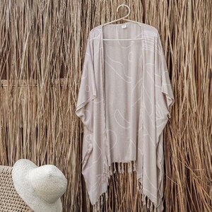 Peut inclure: Un kimono beige avec un motif abstrait blanc, suspendu à un cintre blanc. Le vêtement a des franges sur le bord inférieur. Un chapeau de paille repose sur une chaise en osier à gauche.