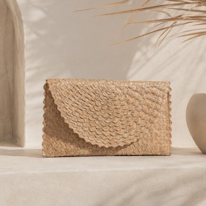 May include: A rectangular clutch bag crafted from woven tan natural fibers. The bag features a distinct envelope-style flap with a rounded, scalloped edge and a textured, layered scale-like pattern across the entire surface of the exterior.