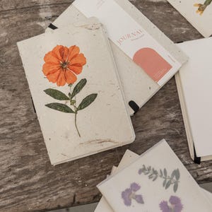 May include: A collection of journals with textured off-white covers. One journal features a pressed orange flower with green leaves. Another journal has a paper band labelled JOURNAL ARTISAN PAPER. A stack of translucent paper sheets displays purple flowers.
