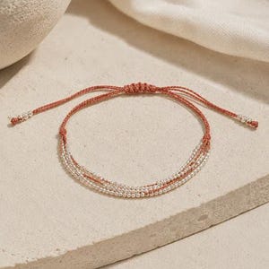 May include: A multi-strand bracelet featuring thin, rust-colored cord intertwined with small, metallic silver beads. The design includes an adjustable sliding knot closure and two dangling cord ends, each finished with a single silver bead at the tip.