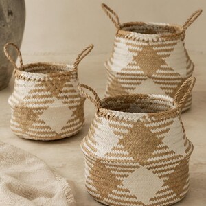 May include: Three woven baskets featuring a geometric diamond pattern in natural tan and white. Each basket has a round base, a tapered middle, and two braided handles attached to the top rim. The baskets are arranged in a cluster on a flat surface.