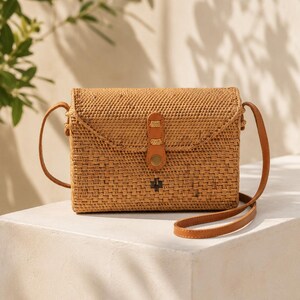 A woven purse with a leather strap sits on a white pedestal against a backdrop of greenery.