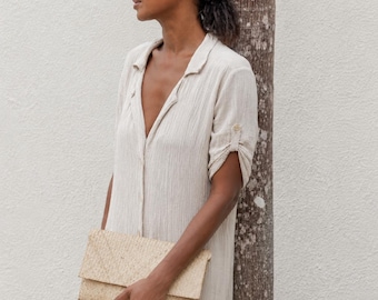 May include: A beige button-down midi dress with a textured fabric, a collar, and cuffed short sleeves, paired with a patterned headband and a rectangular woven straw clutch bag held in front of the body.