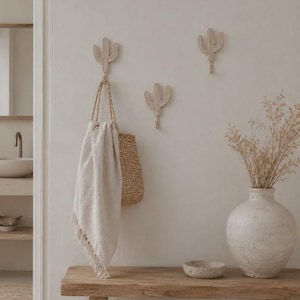 May include: Three light beige cactus-shaped wall hooks made of coiled rope. One hook holds a woven straw bag with a light-colored fabric towel draped over the handle. The hooks are mounted on a plain wall above a wooden bench with a vase of dried flowers.