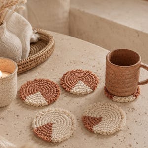 May include: Four circular macrame coasters featuring a spiral pattern in terracotta and cream cotton cord. Each coaster has a distinct color-blocked design with a fringed edge. One coaster is placed under a speckled brown ceramic mug.
