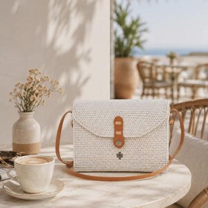 Op de afbeelding: Een rechthoekige crossbodytas van gevlochten rotan in een lichte off-white kleur. De tas heeft een beige leren schouderband en een bijpassende leren sluiting met een klein metalen accent. Het vlechtwerk is strak en gelijkmatig over het oppervlak.