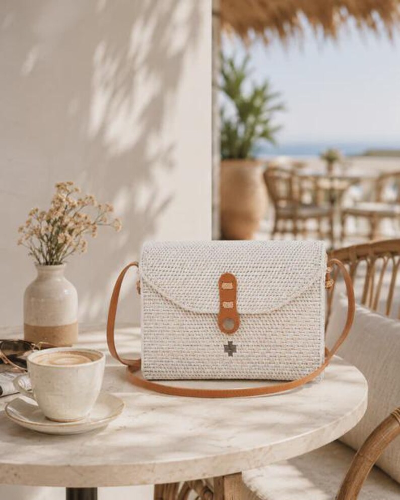 Op de afbeelding: Een rechthoekige crossbodytas van gevlochten rotan in een lichte off-white kleur. De tas heeft een beige leren schouderband en een bijpassende leren sluiting met een klein metalen accent. Het vlechtwerk is strak en gelijkmatig over het oppervlak.